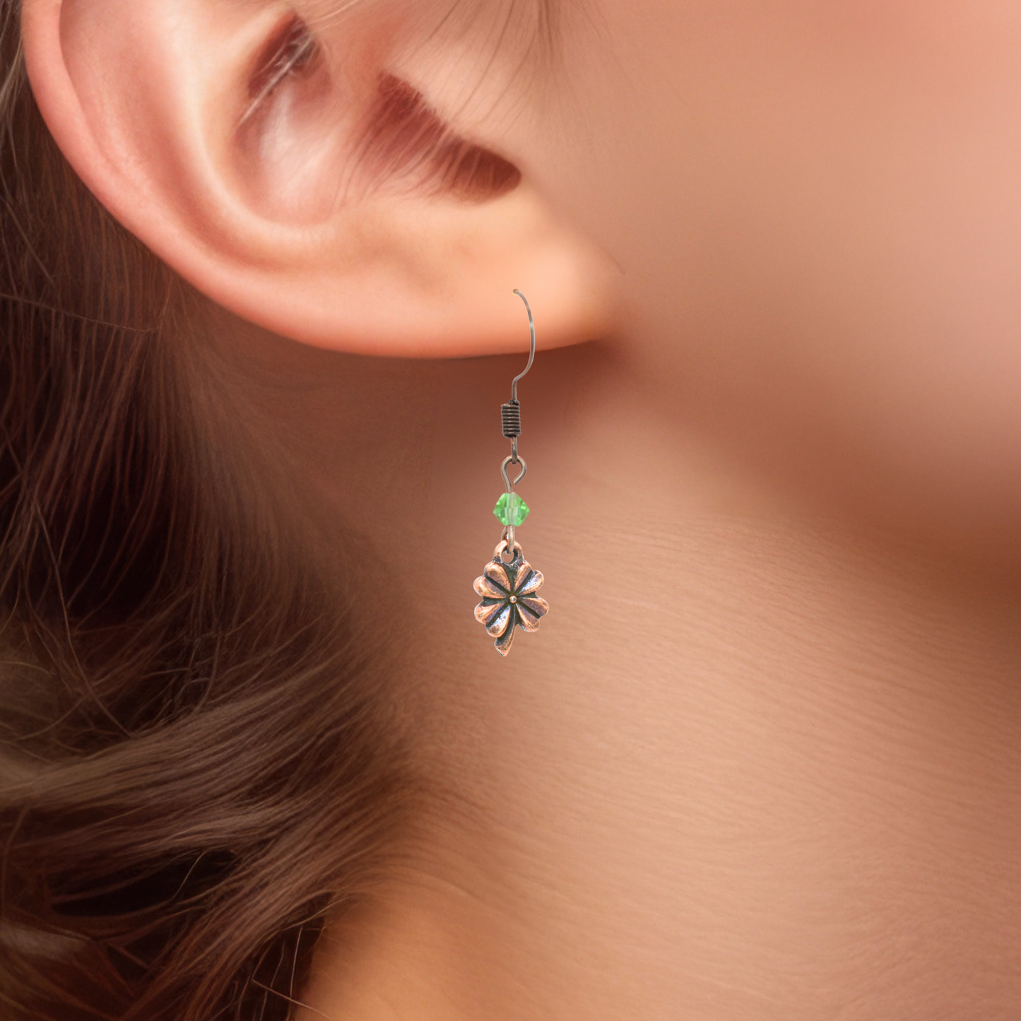 Close-up of an ear wearing an earring featuring a green crystal bead and an antique copper four leaf clover charm.
