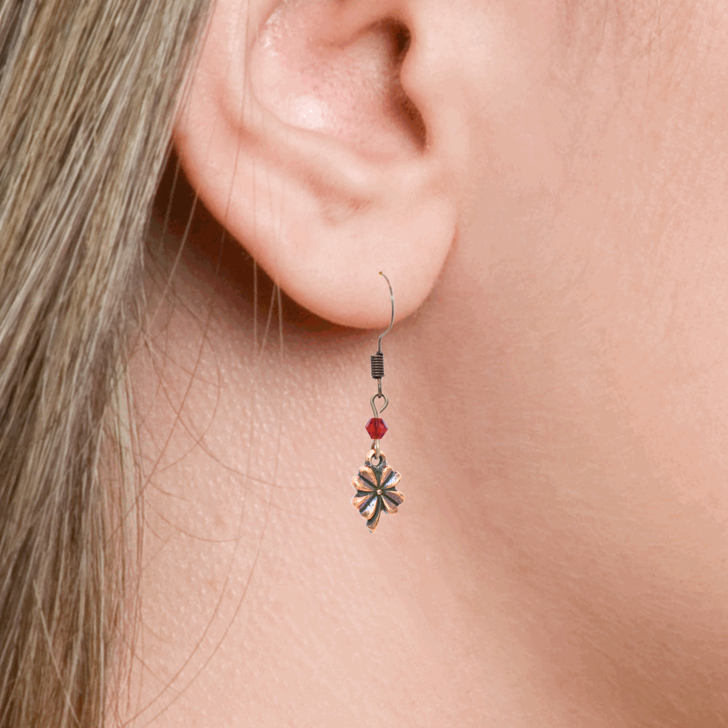 Close-up of an ear wearing an earring featuring a red crystal bead and an antique copper four leaf clover charm.