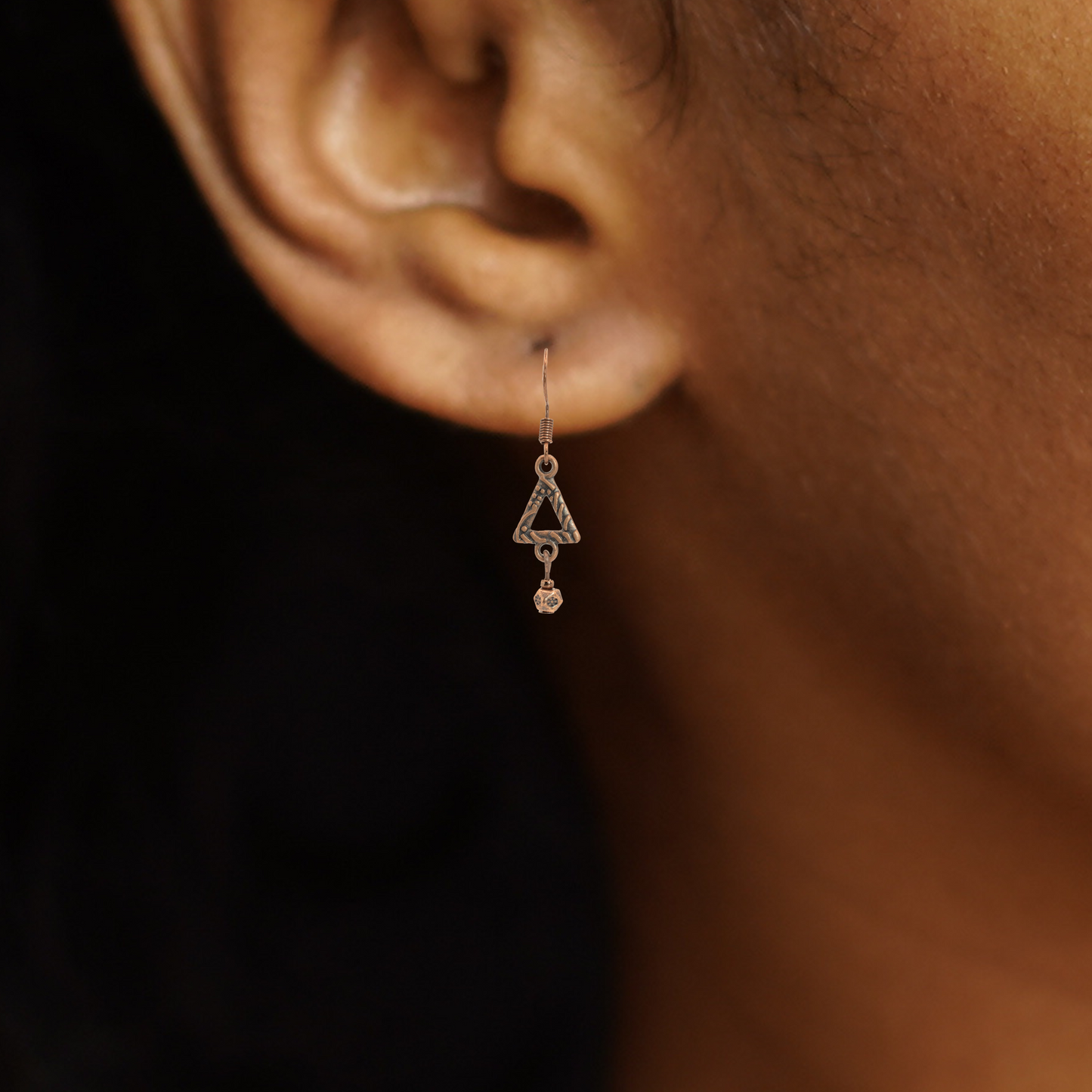 Close-up of a person wearing an antique copper triangle-shaped earring with a dark background