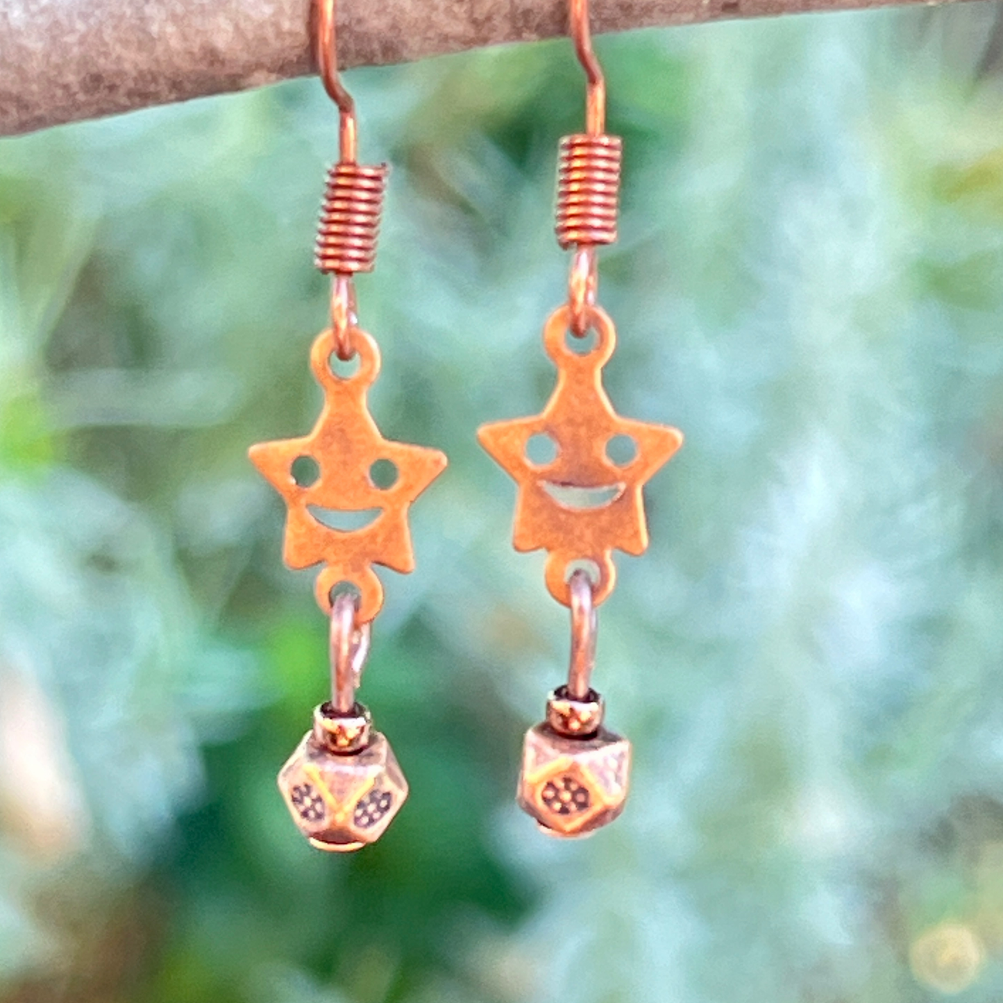 Copper star-shaped earrings with smiley face designs hanging from a branch with green foliage in the background.