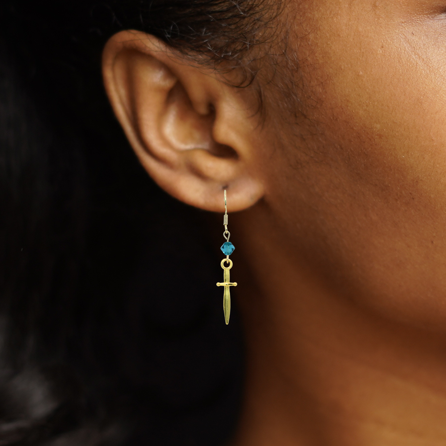 Gold sword earring with blue crystal bead on an ear against a dark background