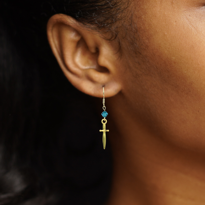 Gold sword earring with blue crystal bead on an ear against a dark background