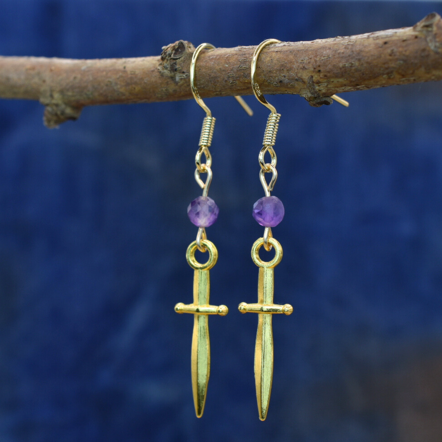 Gold earrings with purple beads hanging on a branch against a blue background