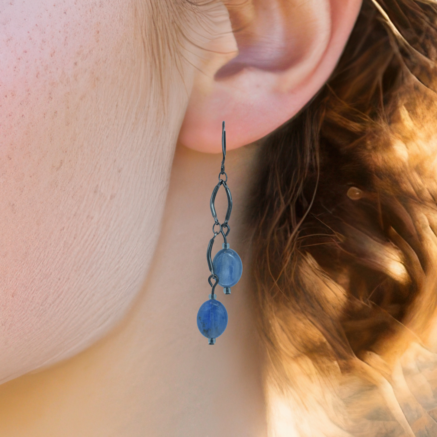 Close-up of an ear wearing a blue dangling earring with blurred hair and skin in the background.