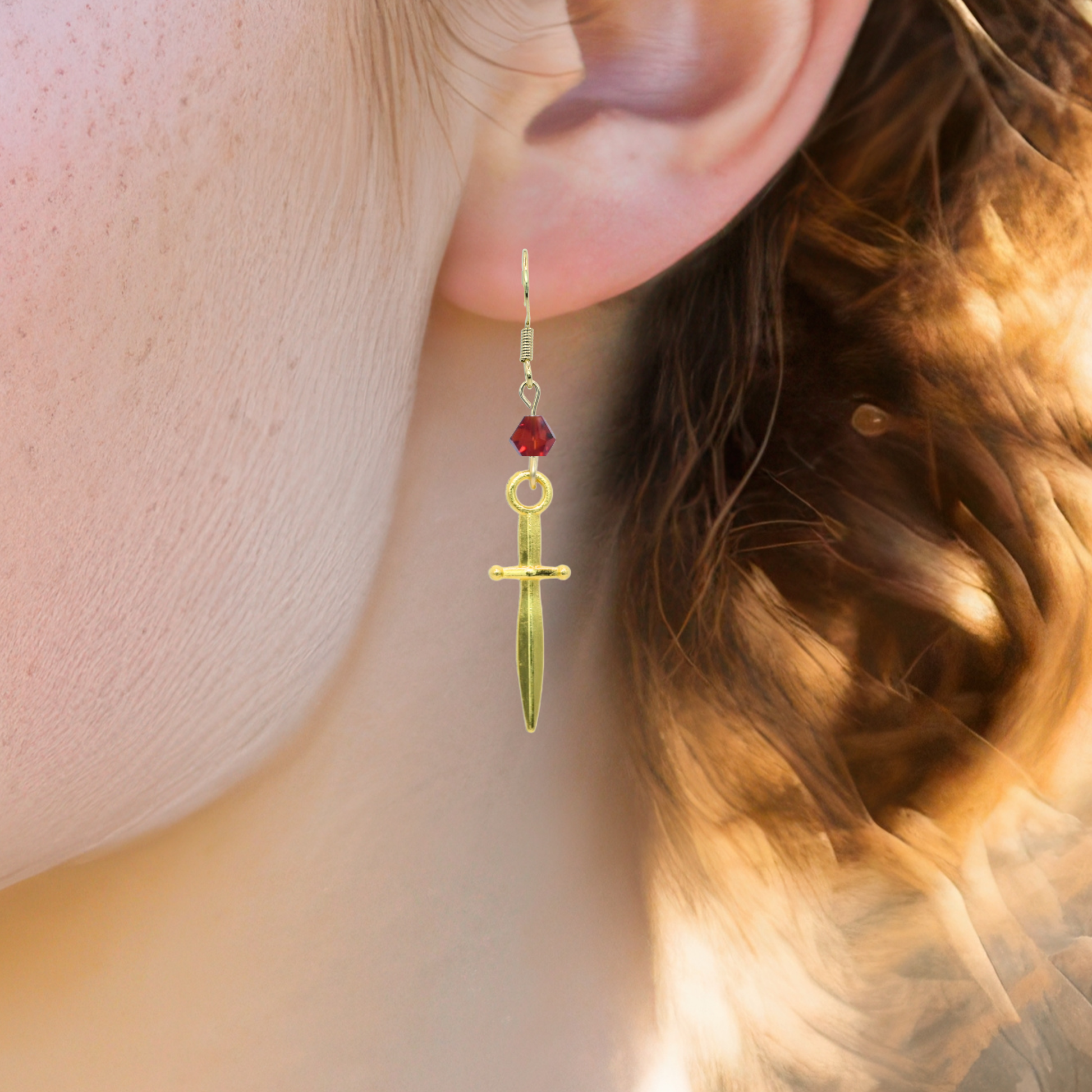 Gold earring with a dagger charm and a red crystal bead worn by a person with brown hair.