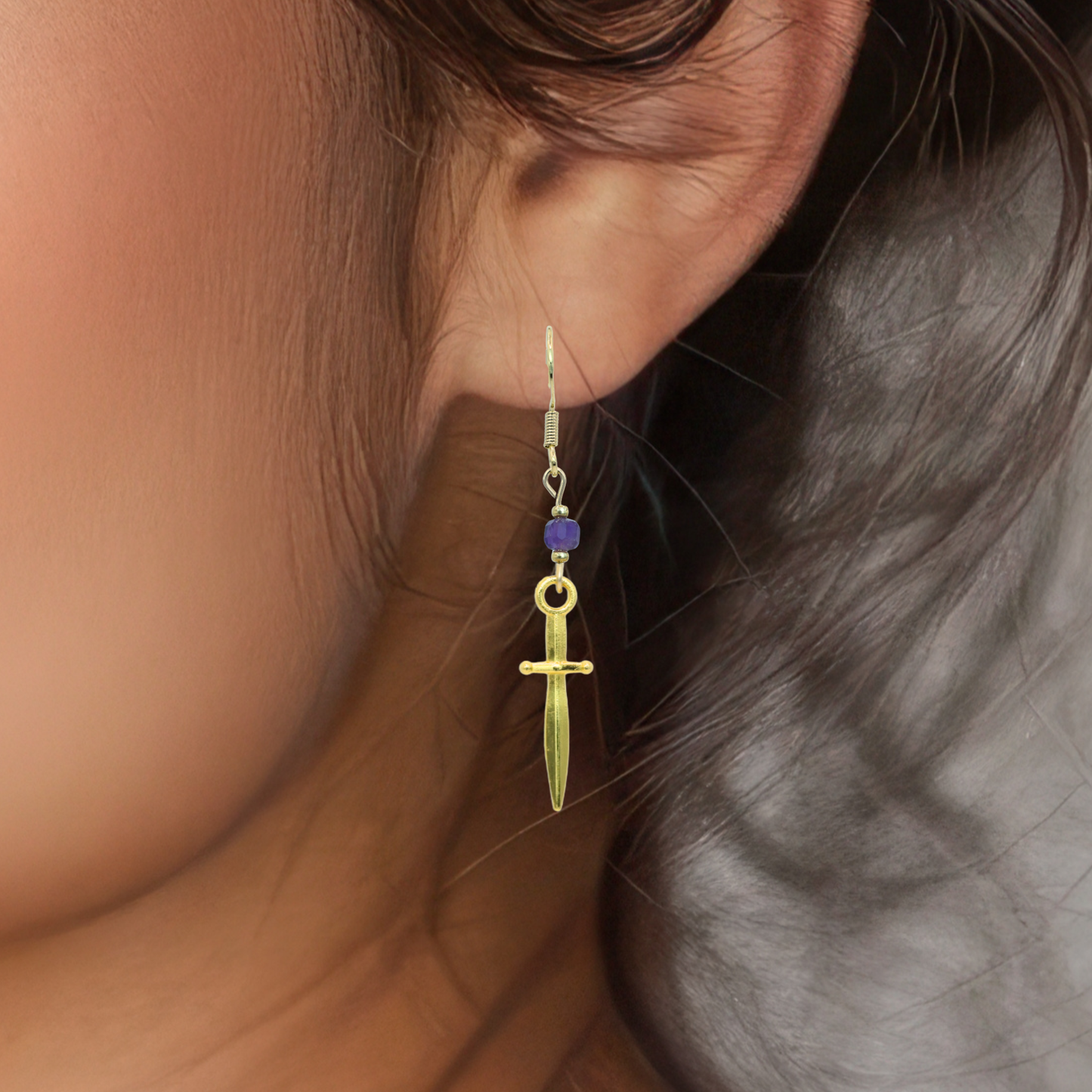 Gold dagger-shaped earring with a purple bead worn by a person.