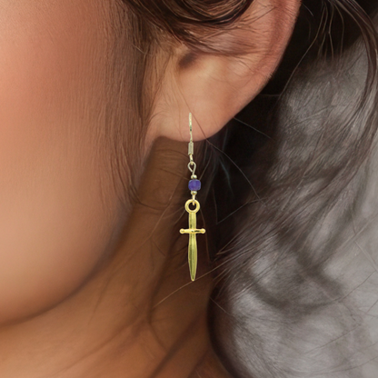 Gold dagger-shaped earring with a purple bead worn by a person.