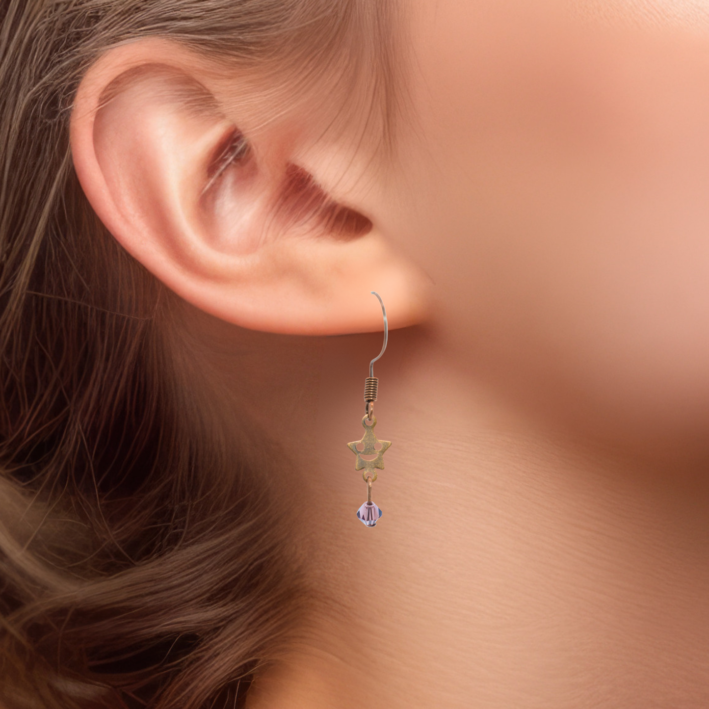 Star-shaped smiley face charm with a light purple crystal bead earring worn on an ear with a soft background