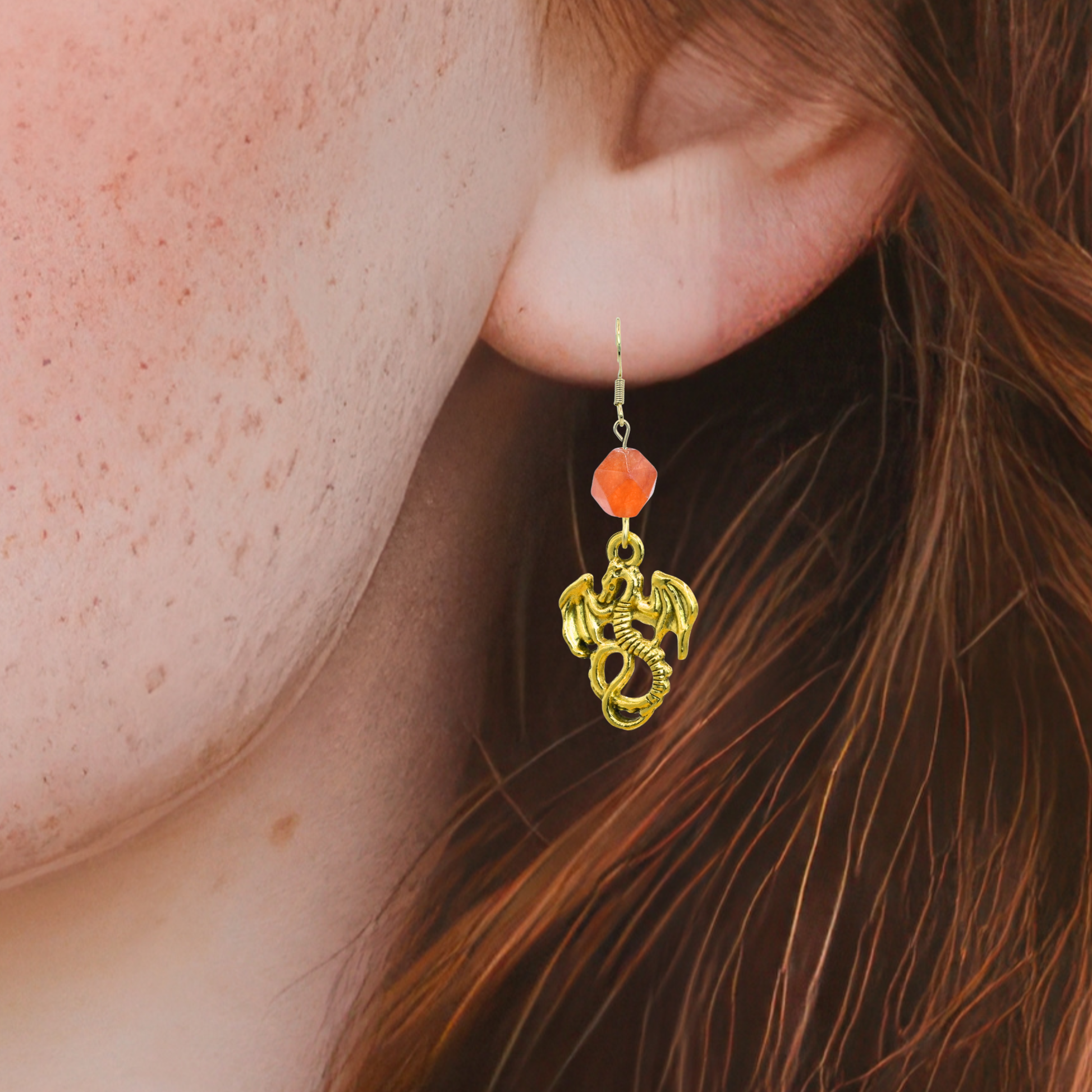 Close-up of a person wearing a gold dragon-shaped earring with an orange bead.